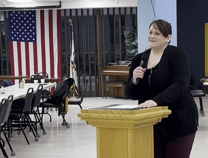 Executive Director Stacie Weaver talks at a podium into a microphone in a room with tables and chairs, with an american flag in the background.