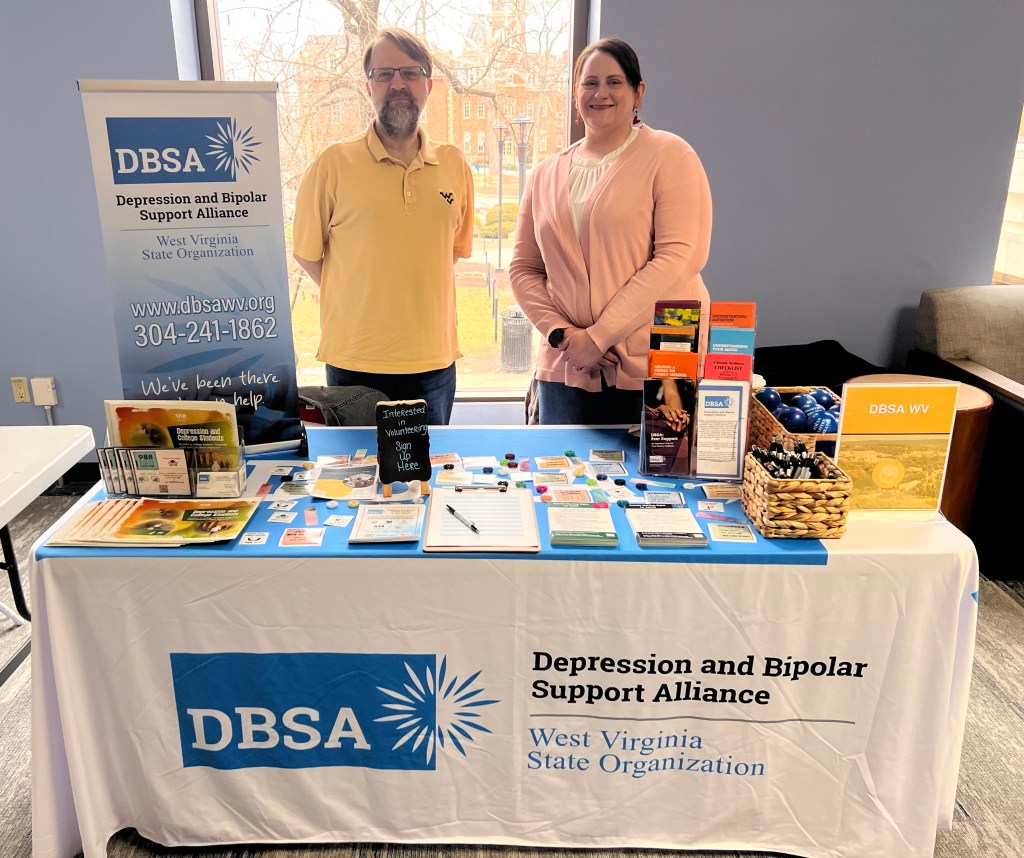 Roger Neptune and Stacie Weaver stand behind the DBSA WV exhibitor table. Signs, flyers, brochures, stress balls, and pens are shown. Table cloth reads "Depression and Bipolar Support Alliance", "West Virginia State Organization".