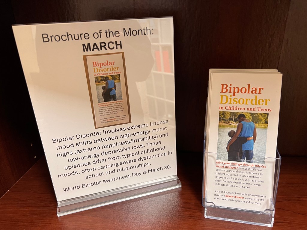 A stack of brochures beside a sign saying "Brochure of the Month: March. Bipolar Disorder involves extreme intense mood shifts between high-energy manic highs (extreme happiness/irritability) and low-energy depressive lows. These episodes differ from typical childhood moods, often causing severe dysfunction in school and relationships. World Bipolar Awareness Day is March 30."