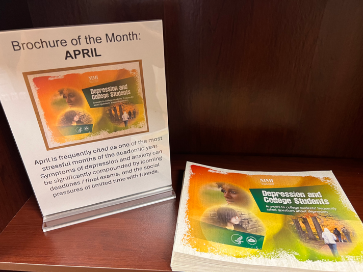 A stack of brochures beside a sign saying "Brochure of the Month: April. April is frequently cited as one of the most stressful months of the academic year. Symptoms of depression and anxiety can be significantly compounded by looming deadlines / final exams, and the social pressures of limited time with friends."