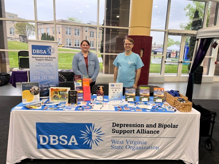 Stacie Weaver and Roger Neptune stand behind the DBSA WV exhibitor table. Signs, flyers, brochures, stress balls, pens and stickers are shown on the table. Table cloth reads "DBSA Depression and Bipolar Support Alliance", "West Virginia State Organization".