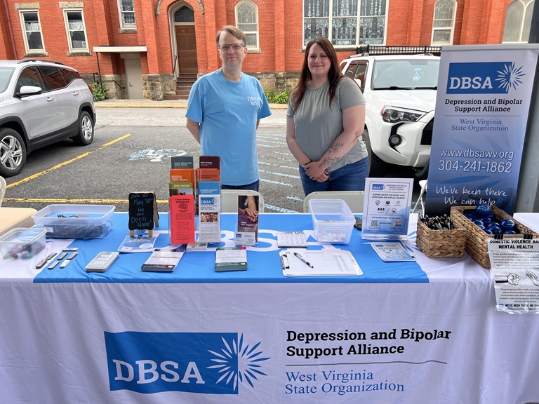 Roger Neptune and Stacie Weaver stand behind the DBSA WV exhibitor table. Signs, flyers, brochures, stress balls, and pens are shown. Table cloth reads "Depression and Bipolar Support Alliance", "West Virginia State Organization".