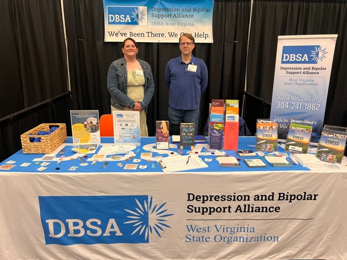 Stacie Weaver and Roger Neptune stand behind the DBSA WV exhibitor table. Signs, flyers, brochures, stress balls, and stickers are shown on the table. Table cloth reads "DBSA Depression and Bipolar Support Alliance", "West Virginia State Organization".