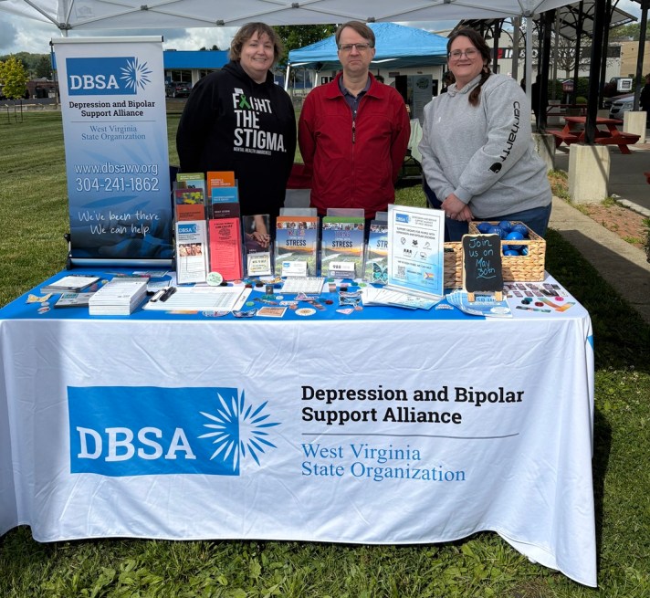 Kari Williams, Roger Neptune, and Stacie Weaver stand behind the DBSA WV exhibitor table. Signs, flyers, brochures, stress balls, and stickers are shown on the table. Table cloth reads "DBSA Depression and Bipolar Support Alliance", "West Virginia State Organization".