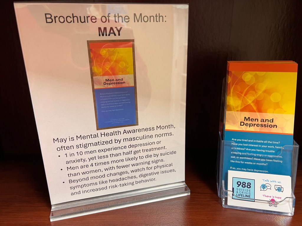 A stack of brochures beside a sign saying "Brochure of the Month: May. May is Mental Health Awareness Month, often stigmatized by masculine norms. 
1 in 10 men experience depression or anxiety, yet less than half get treatment.
Men are 4 times more likely to die by suicide than women, with fewer warning signs. 
Beyond mood changes, watch for physical symptoms like headaches, digestive issues, and increased risk-taking behavior."