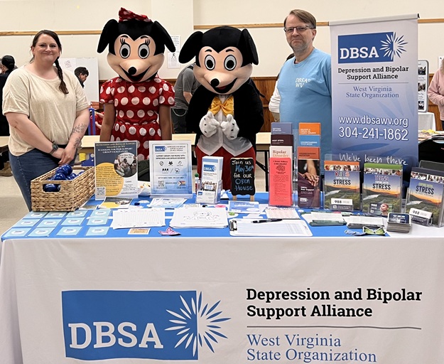 Stacie Weaver and Roger Neptune stand behind the DBSA WV exhibitor table along with 2 people in costume looking like Mickey and Minnie Mouse.