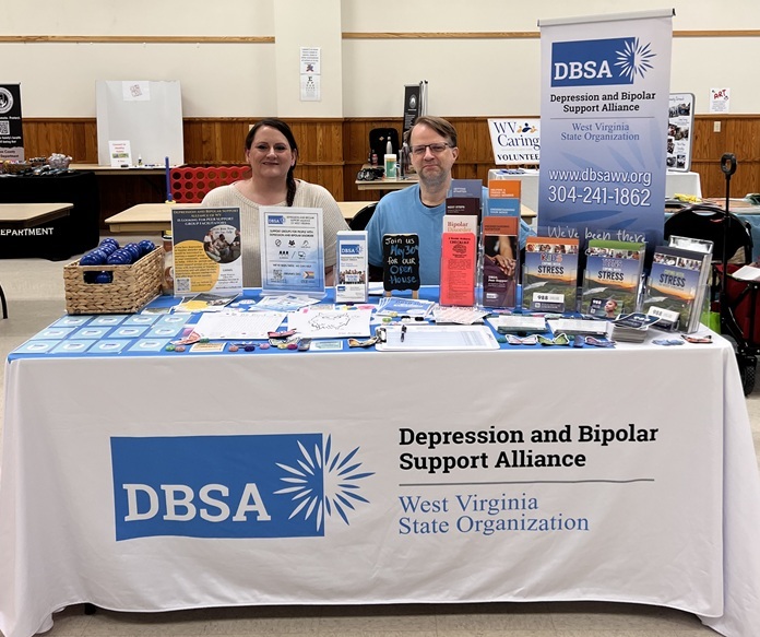 Stacie Weaver and Roger Neptune sit behind the DBSA WV exhibitor table. Signs, flyers, brochures, stress balls, and stickers are shown on the table. Table cloth reads "DBSA Depression and Bipolar Support Alliance", "West Virginia State Organization".