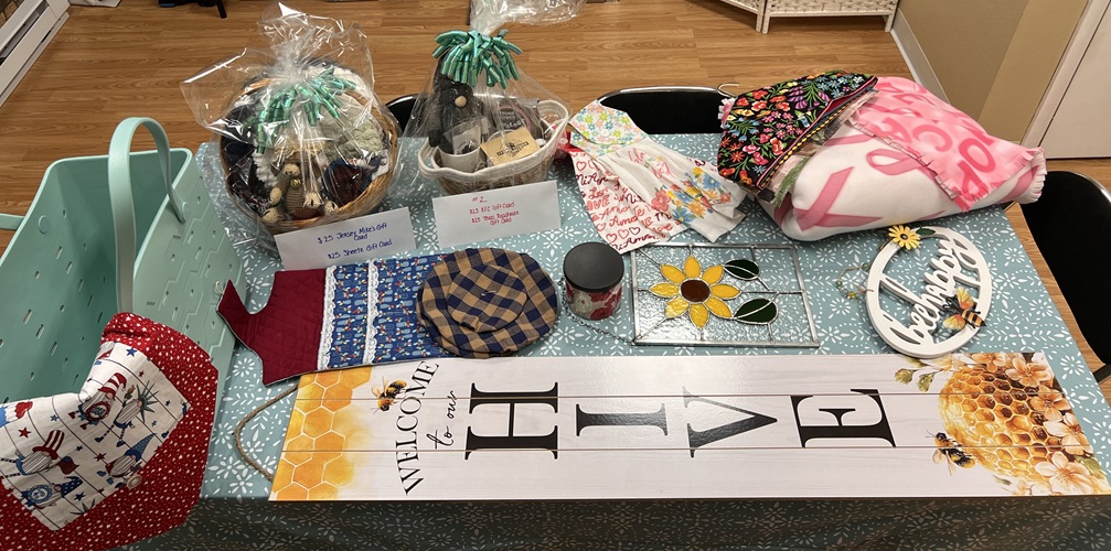 A table full of items, such as gift cards, gift baskets, a blanket, a stained glass window panel, outdoor welcome sign, and more.