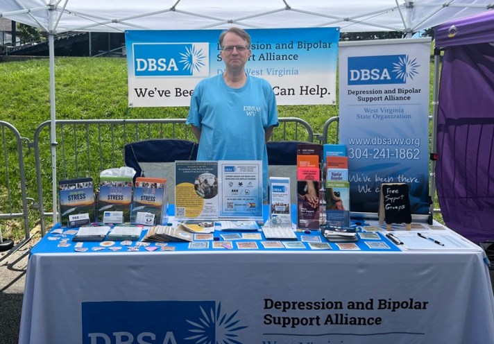 Man standing behind the DBSA WV exhibitor table, under a white canopy, with a DBSA banner behind him. Signs, flyers, brochures, and stickers are shown on the table. Table cloth reads "DBSA Depression and Bipolar Support Alliance".
