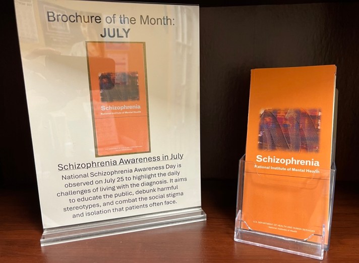 A stack of brochures beside a sign saying "Brochure of the Month: July. Schizophrenia Awareness in July. National Schizophrenia Awareness Day is observed on July 25 to highlight the daily challenges of living with the diagnosis. It aims to educate the public, debunk harmful stereotypes, and combat the social stigma and isolation that patients often face."