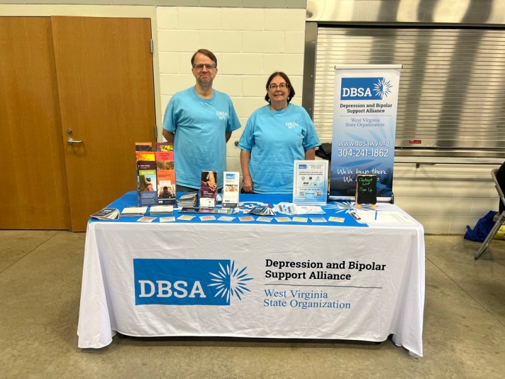 Two people standing behind a table filled with signs, and brochures. The tablecloth reads "DBSA Depression and Bipolar Support Alliance West Virginia State Organization".