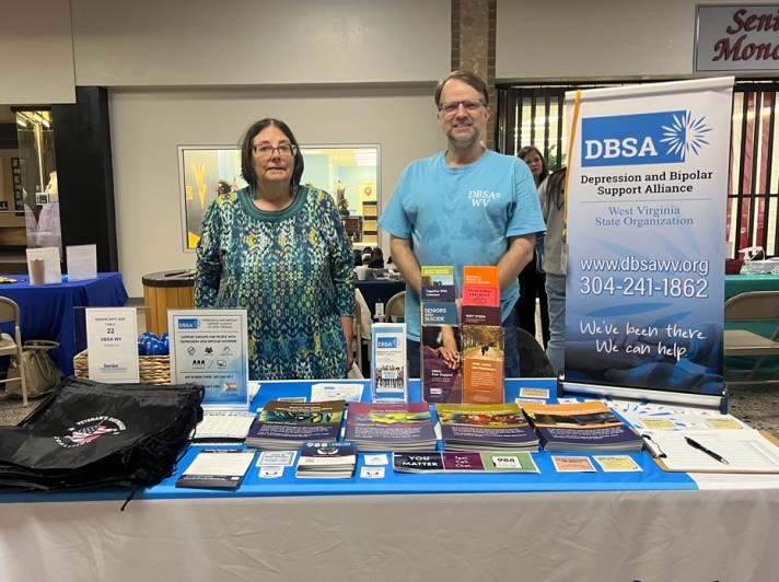 Diane Williams and Roger Neptune stand behind the DBSA WV exhibitor table. Signs, brochures, bags, and stickers are shown on the table.