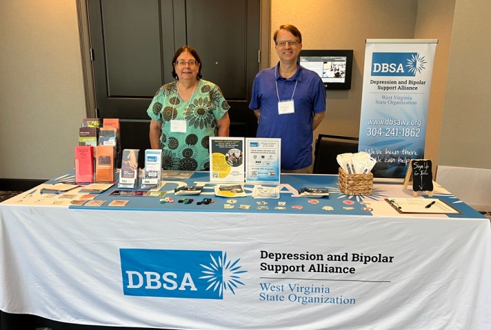 Diane Williams and Roger Neptune stand behind the DBSA WV exhibitor table. Signs, flyers, brochures, pens, and stickers are shown on the table. Table cloth reads "DBSA Depression and Bipolar Support Alliance", "West Virginia State Organization".