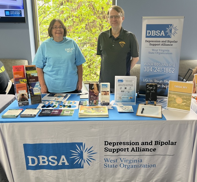 Diane Williams and Roger Neptune stand behind the DBSA WV exhibitor table. Signs, flyers, brochures, and stickers are shown on the table. Table cloth reads "DBSA Depression and Bipolar Support Alliance", "West Virginia State Organization".